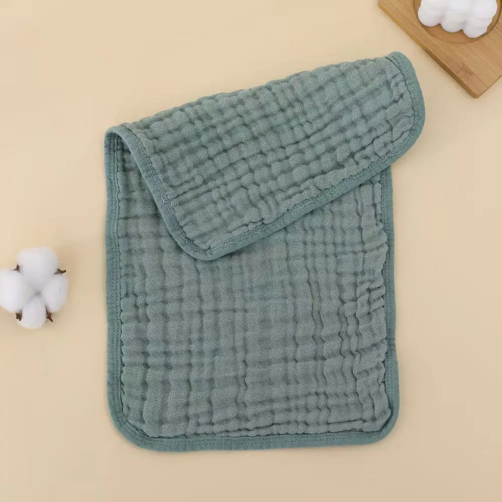 A stack of soft, sage green muslin baby burp cloths with a quilted texture and white cotton balls.
