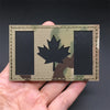 A tactical patch featuring a black and olive green camouflage background with a silver maple leaf emblem.