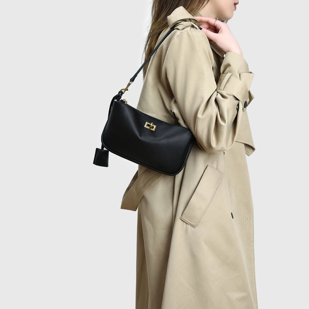 A woman is holding a beige trench coat and a black leather tote bag with a gold metal buckle.