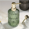 A clear green glass bottle with a gold pump dispenser sits on a white marble surface.