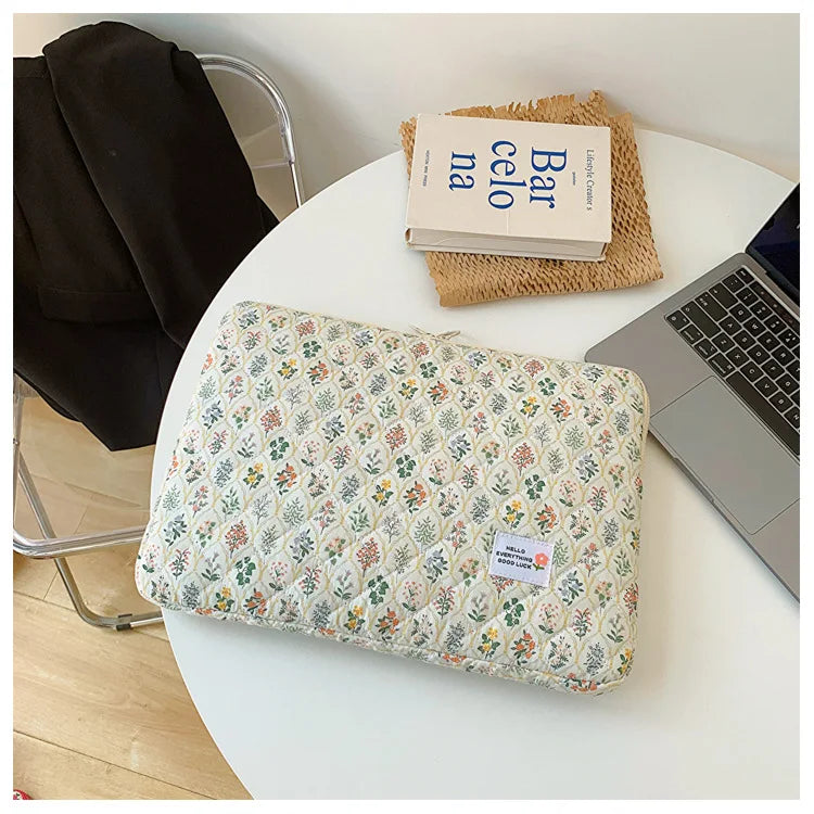 A floral printed laptop sleeve with a zipper closure resting on a white table next to a notebook and a laptop.