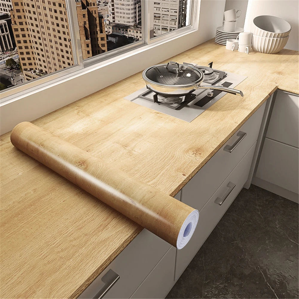A wooden roll of self-adhesive wallpaper is placed on a kitchen countertop next to a window view.