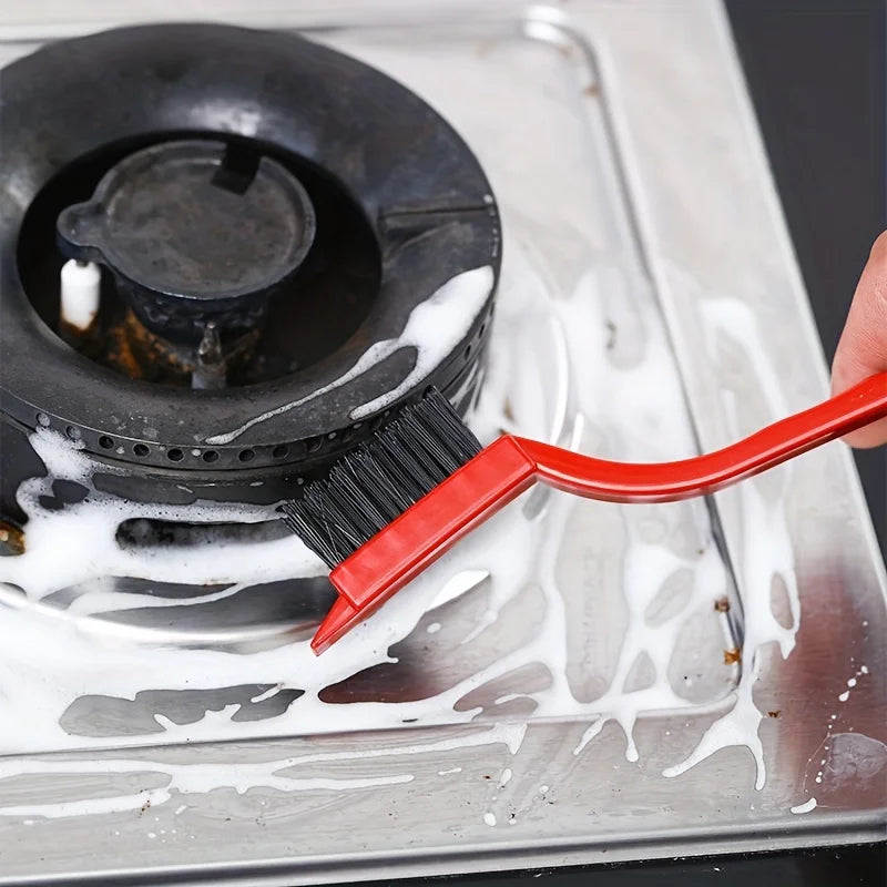 A red kitchen cleaning brush and scraper tool is used to clean food residue from a metal cooking surface.