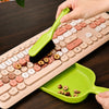 A hand is using a green handheld dustpan and brush set on a beige keyboard, with a green plastic tray nearby.