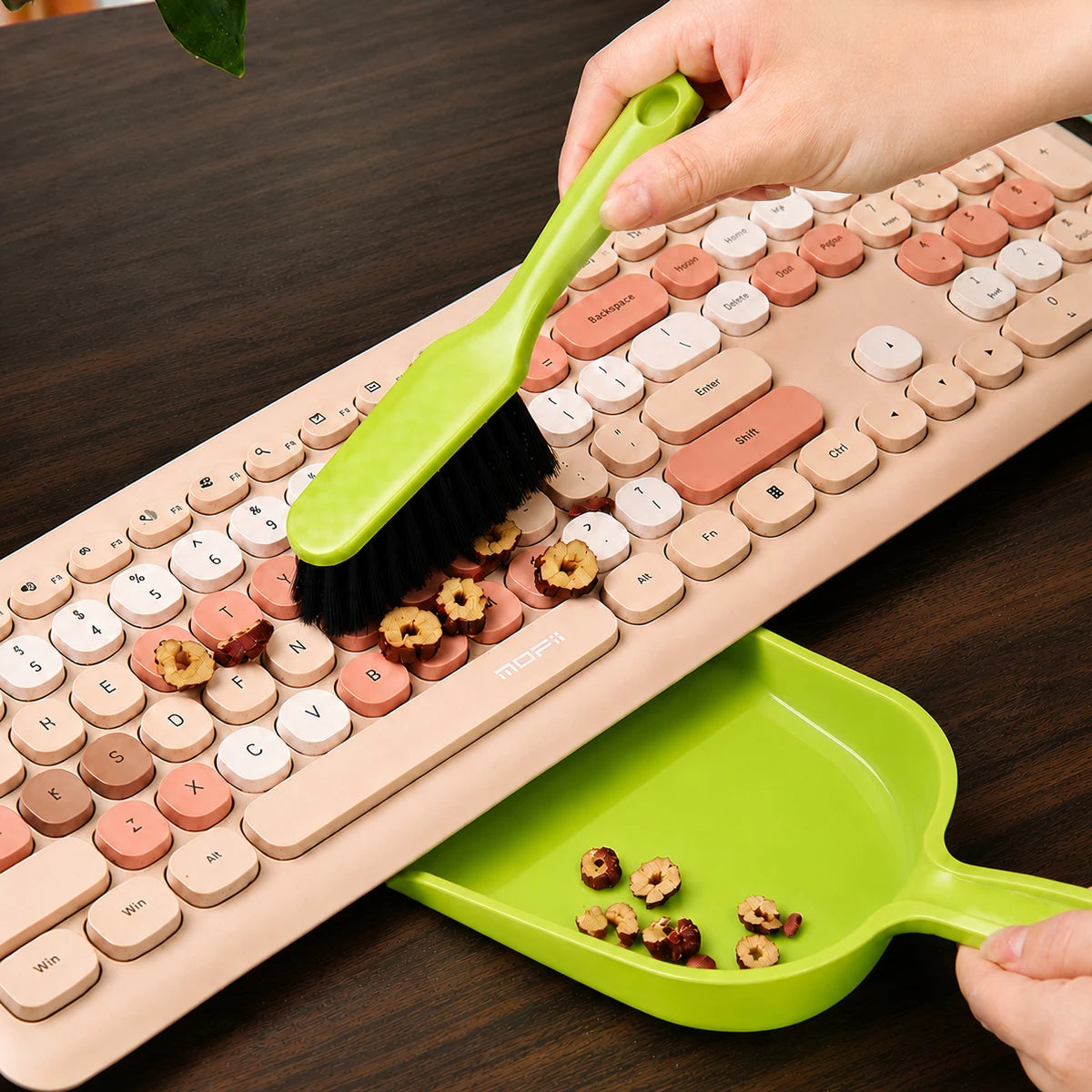 A hand is using a green handheld dustpan and brush set on a beige keyboard, with a green plastic tray nearby.