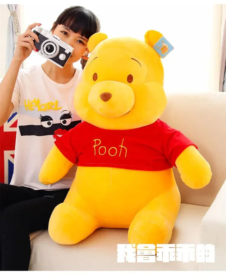 A yellow Winnie the Pooh plush toy sitting on a light-colored sofa, next to a person holding a camera.