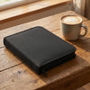A black zippered pouch containing drawing tools like graphite and charcoal pencils rests on a wooden table next to a cup of c