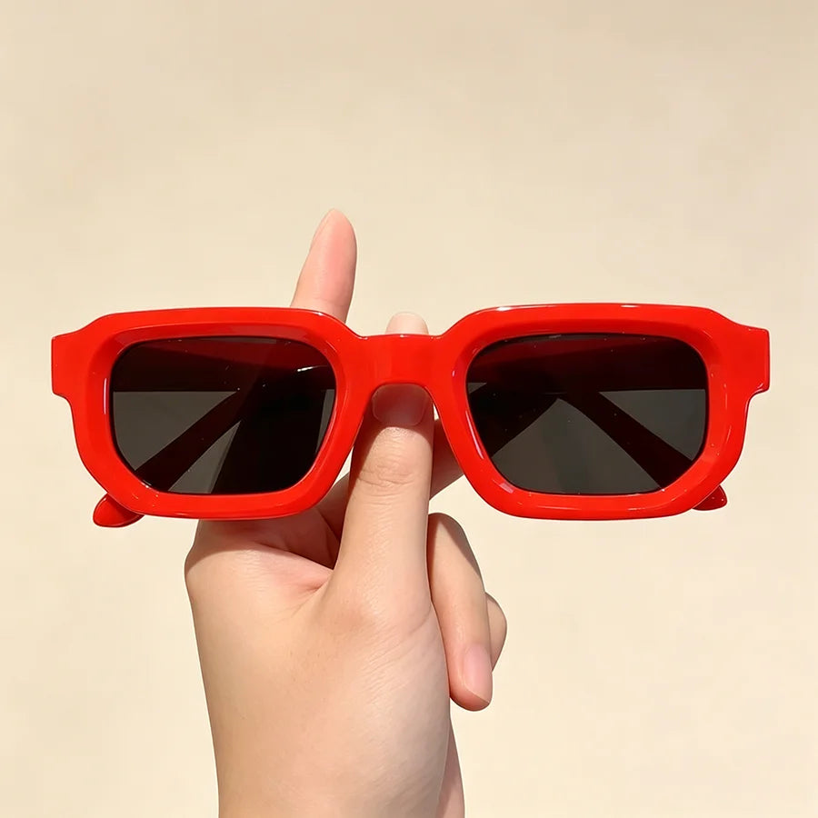 A hand holds a pair of bright red vintage rectangle sunglasses against a light background.