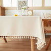 A cream-colored farmhouse tablecloth with delicate cotton tassels draped over a wooden dining table.