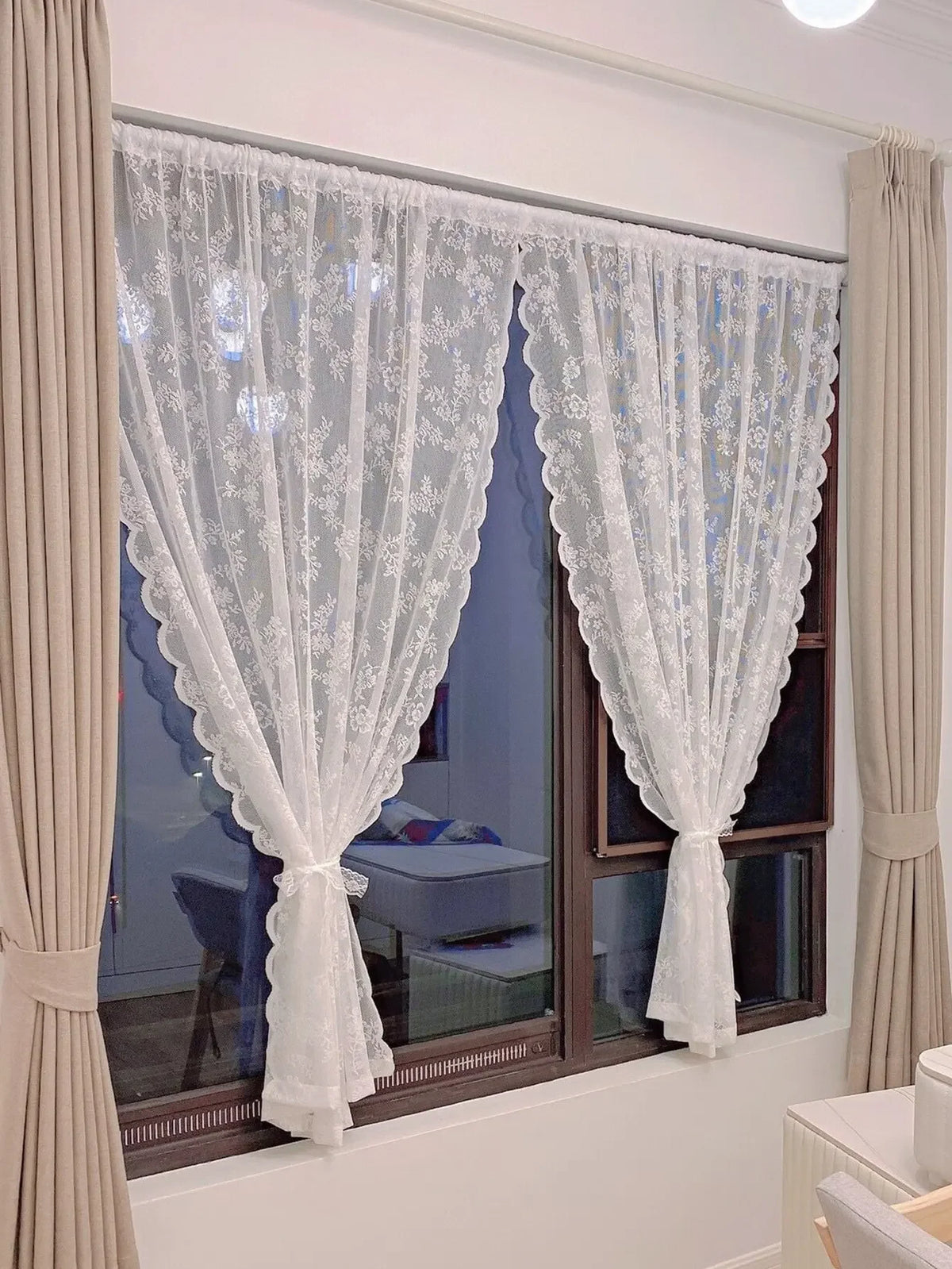 Rideaux transparents avec broderie florale blanche, suspendus à une fenêtre avec des draperies neutres.