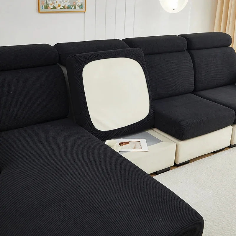 A black Jacquard stretch sofa slipcover with a cream accent cushion shown in a bright room.