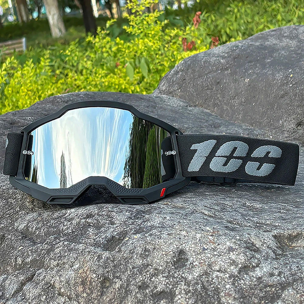A pair of black motocross goggles with a reflective lens and a black strap resting on a large gray rock.