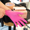 A hand wearing a bright pink nitrile disposable glove is holding a box of pink cosmetic products.
