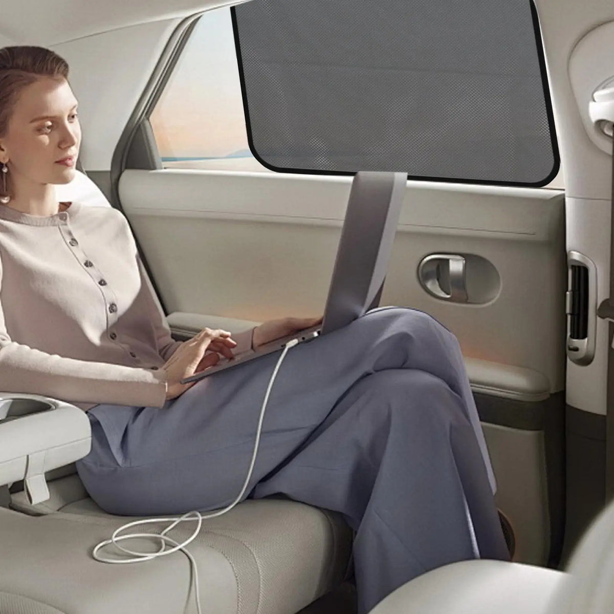 A woman sits in a car, using a laptop while magnetic car window sunshades are visible.