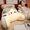 A light gray plush cat and dog backrest cushion is shown resting on a brown sofa, with a woman nearby.