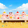 Four pink resin pig figurines with colorful accessories sitting on a wooden surface against a bright blue sky and yellow sand