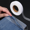 A hand is holding a roll of white double-sided iron-on adhesive tape next to a piece of denim fabric.