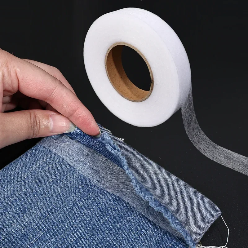 A hand is holding a roll of white double-sided iron-on adhesive tape next to a piece of denim fabric.