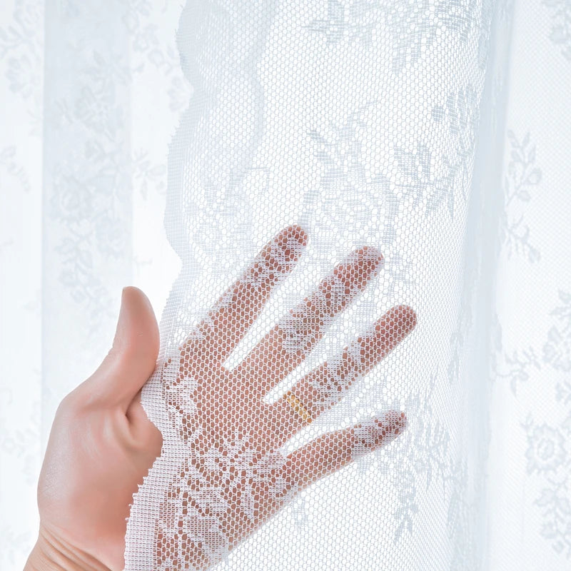 A hand is holding a sheer white floral lace curtain panel, showing the intricate texture and light filtering through.