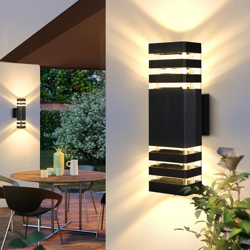 A modern black LED wall sconce with vertical light strips illuminates a white wall outdoors.