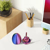 A colorful, iridescent middle finger egg sits on a white desk next to pencils and a plant.