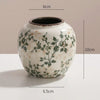 Weiße Keramikvase mit grünen Blumenmustern und dunklem Rand, vor hellem Hintergrund.