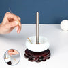 A hand holds a curved stainless steel beading needle above a white bowl and a dark wooden base.