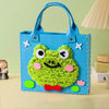 A bright blue felt tote bag shaped like a smiling green frog with large eyes and a red bow.