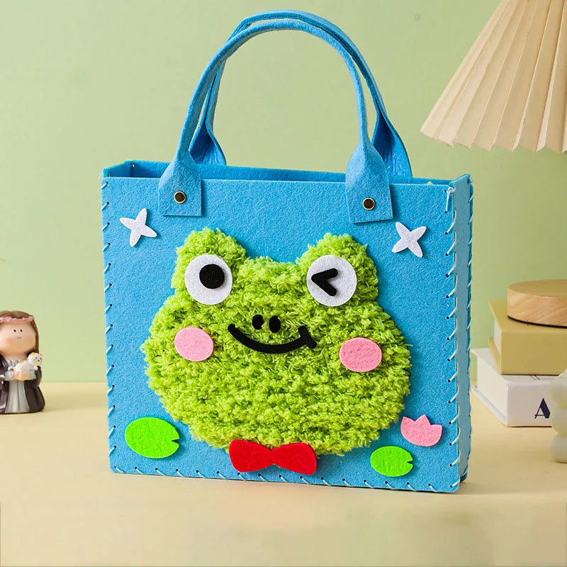 A bright blue felt tote bag shaped like a smiling green frog with large eyes and a red bow.
