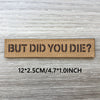 A rectangular brown woven patch with the text BUT DID YOU DIE? on a light wooden surface.