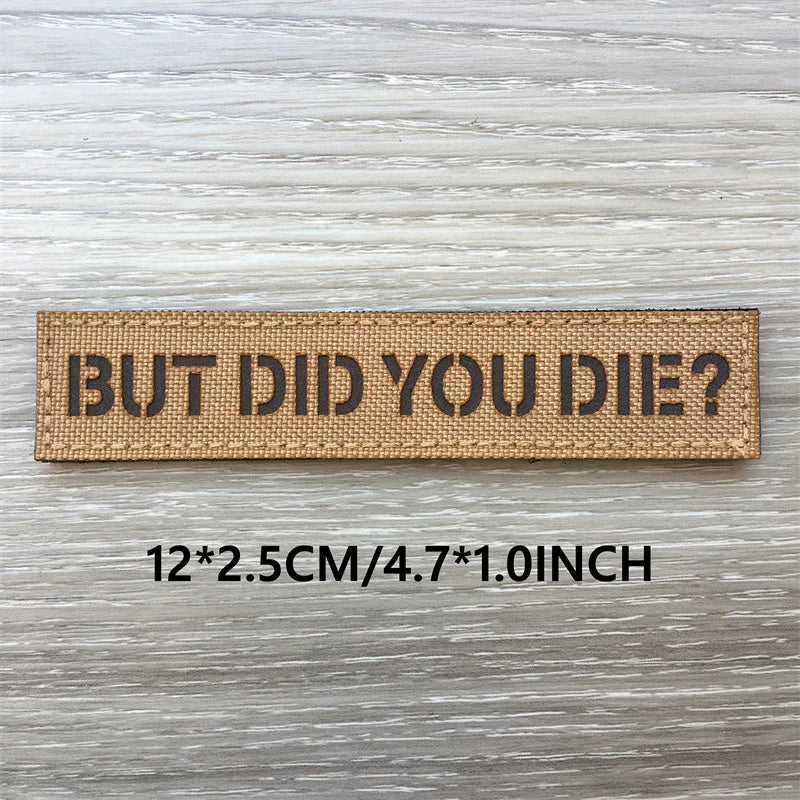 A rectangular brown woven patch with the text BUT DID YOU DIE? on a light wooden surface.