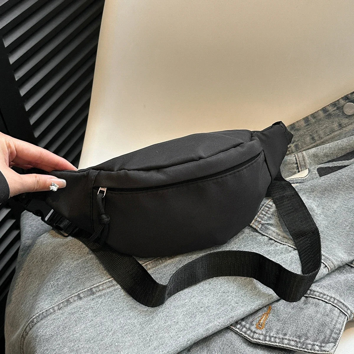 Una mano sostiene un bolso cruz negro de nylon contra una camisa vaquera gris.