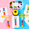 A set of Montessori wooden bead sorting game components including a clear plastic box, yellow bead, and test tubes with color