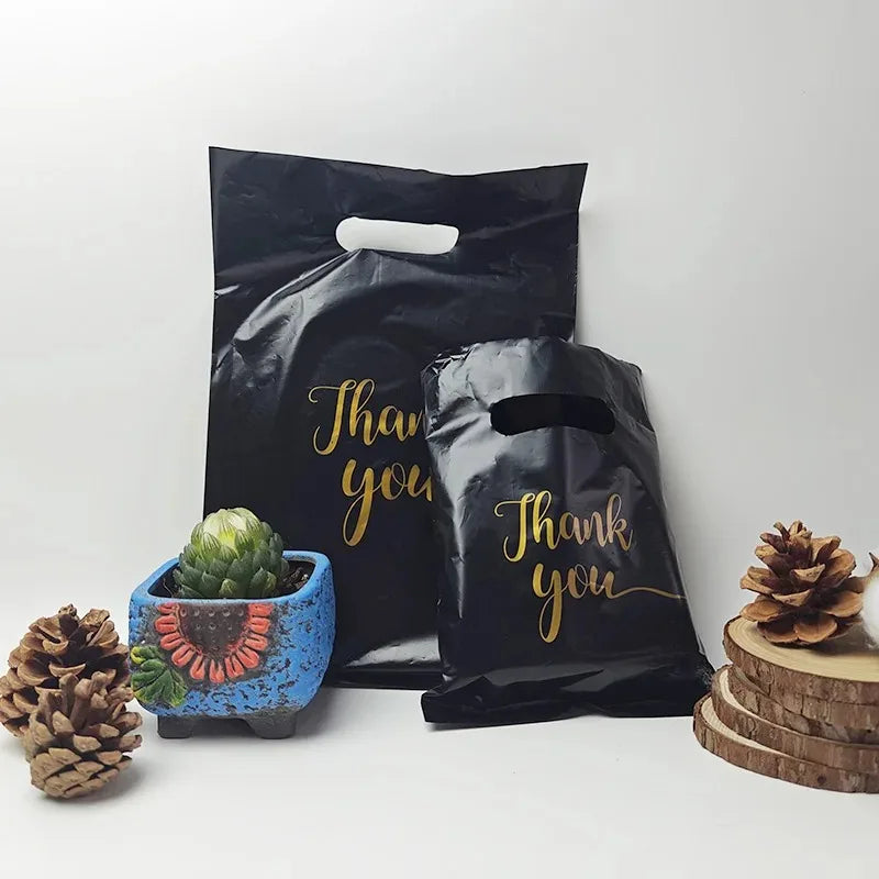 A set of black thank you gift bags with gold lettering and natural elements like pinecones and succulents.