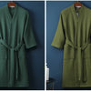 A long, olive green waffle knit robe hanging on a wooden hanger against a dark blue wall.