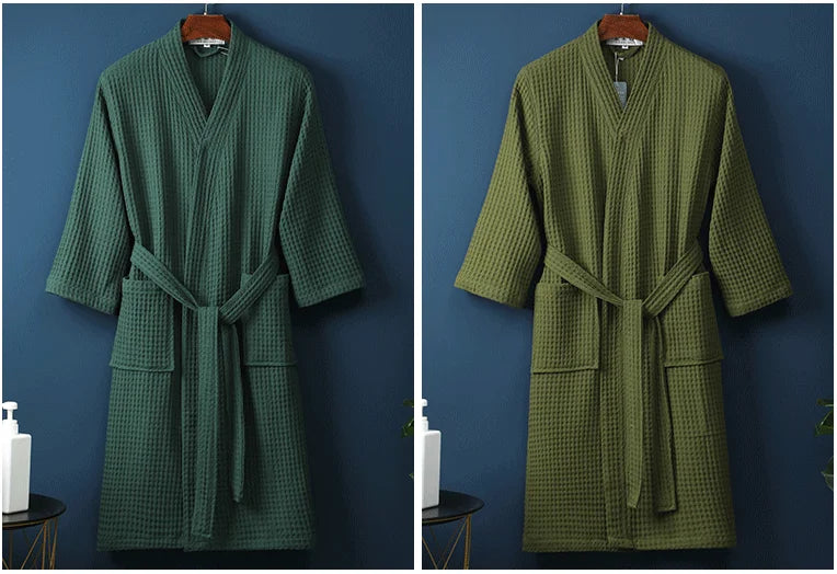 A long, olive green waffle knit robe hanging on a wooden hanger against a dark blue wall.