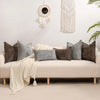 A collection of gray and brown textured square throw pillows arranged on a white sofa with a cream blanket and natural light 