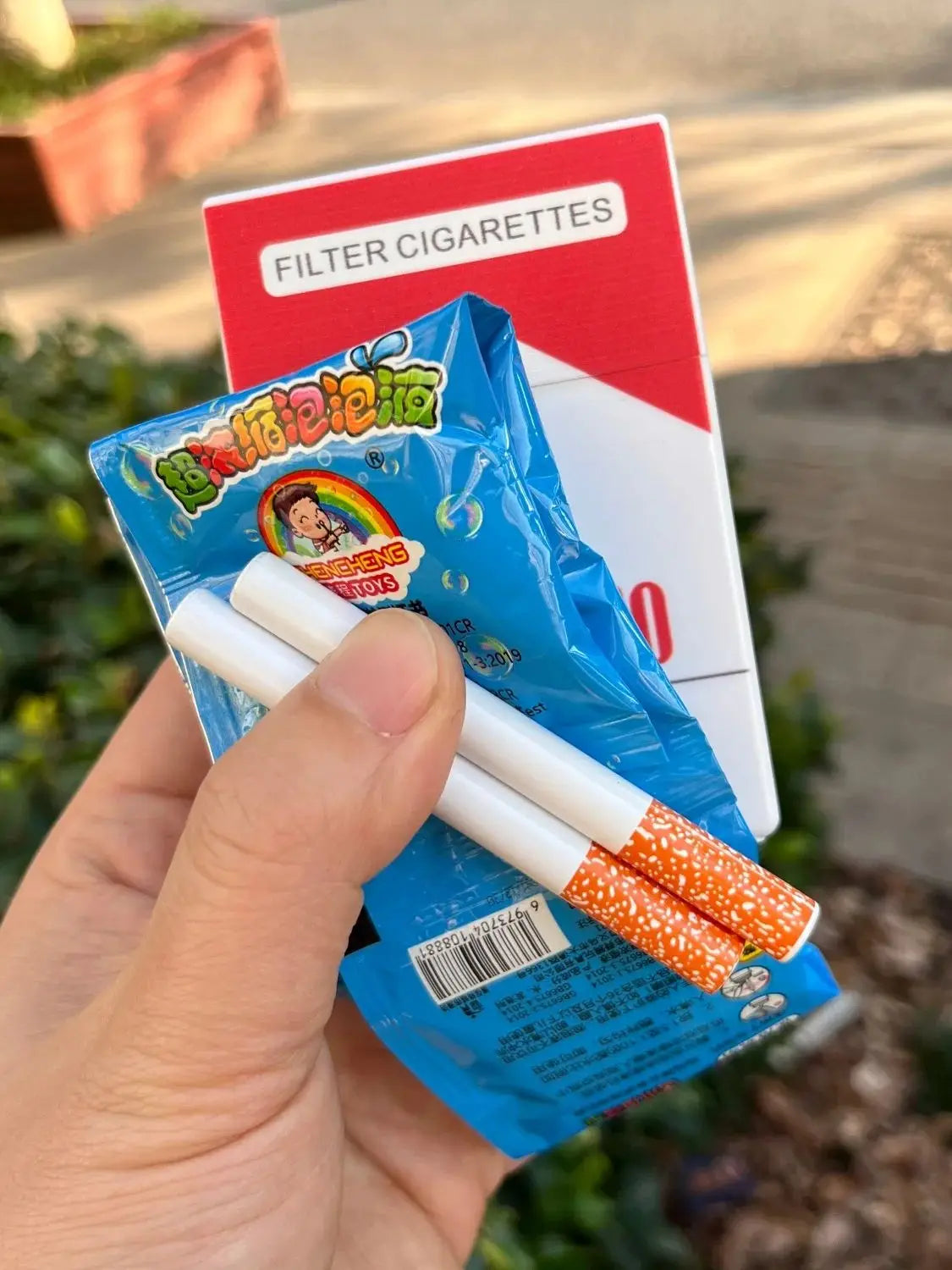 A hand holds a blue packet of funny bubble blowing pipes and a red card labeled Filter Cigarettes.