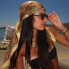 A woman wearing a leopard paisley square scarf and sunglasses stands near the sea.