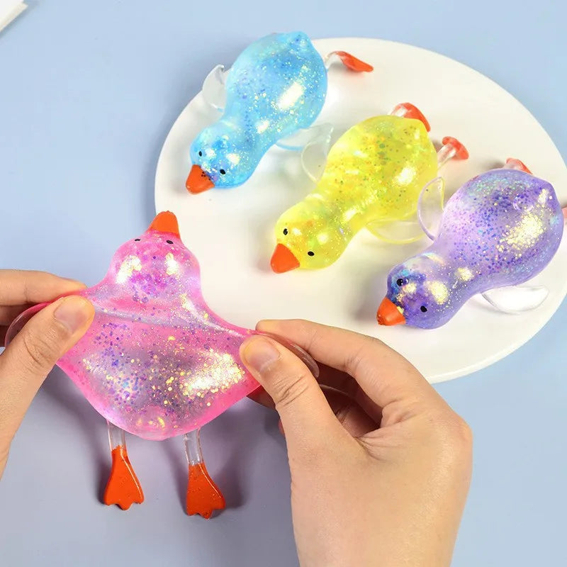 Hände drücken einen rosa Glitzer-Enten-Stressball neben zwei gelb-blauen Glitzerenten auf einer weißen Platte.