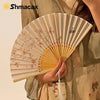 A close-up of a light beige bamboo and silk folding fan with delicate pink floral designs being held.