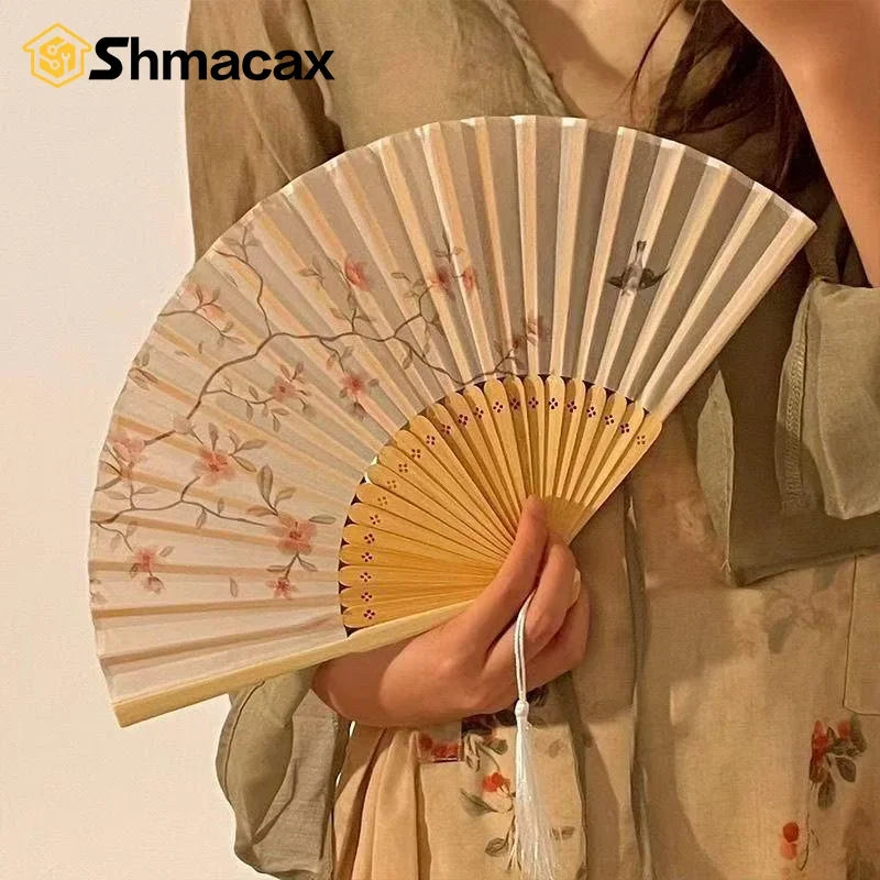 A close-up of a light beige bamboo and silk folding fan with delicate pink floral designs being held.