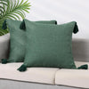 A dark green cotton linen throw pillow with tassels resting on a light colored sofa.