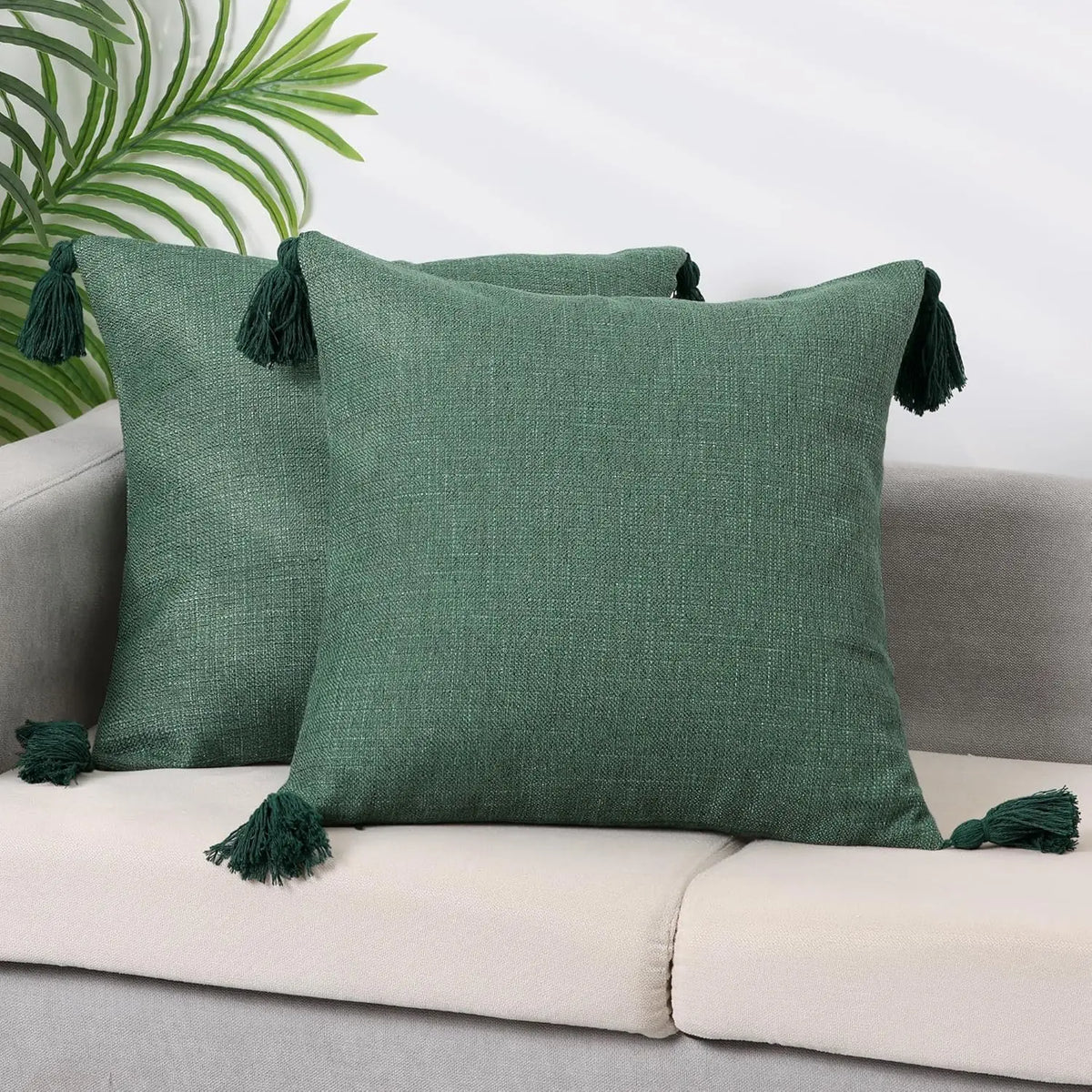 A dark green cotton linen throw pillow with tassels resting on a light colored sofa.