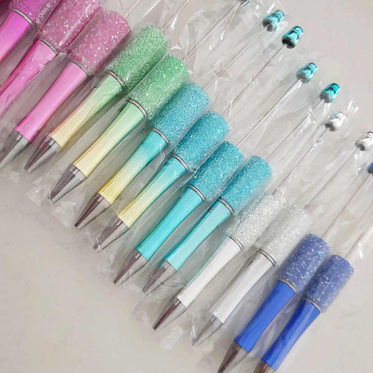 Collection de 30 stylos-plumes à bille en perles dans diverses nuances de rose, vert et bleu, présentés dans un sac plastique
