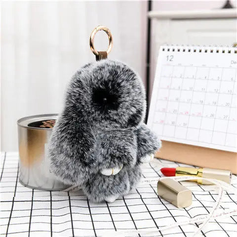 A fluffy gray bunny keychain with a black pom pom tail resting on a black and white grid surface.