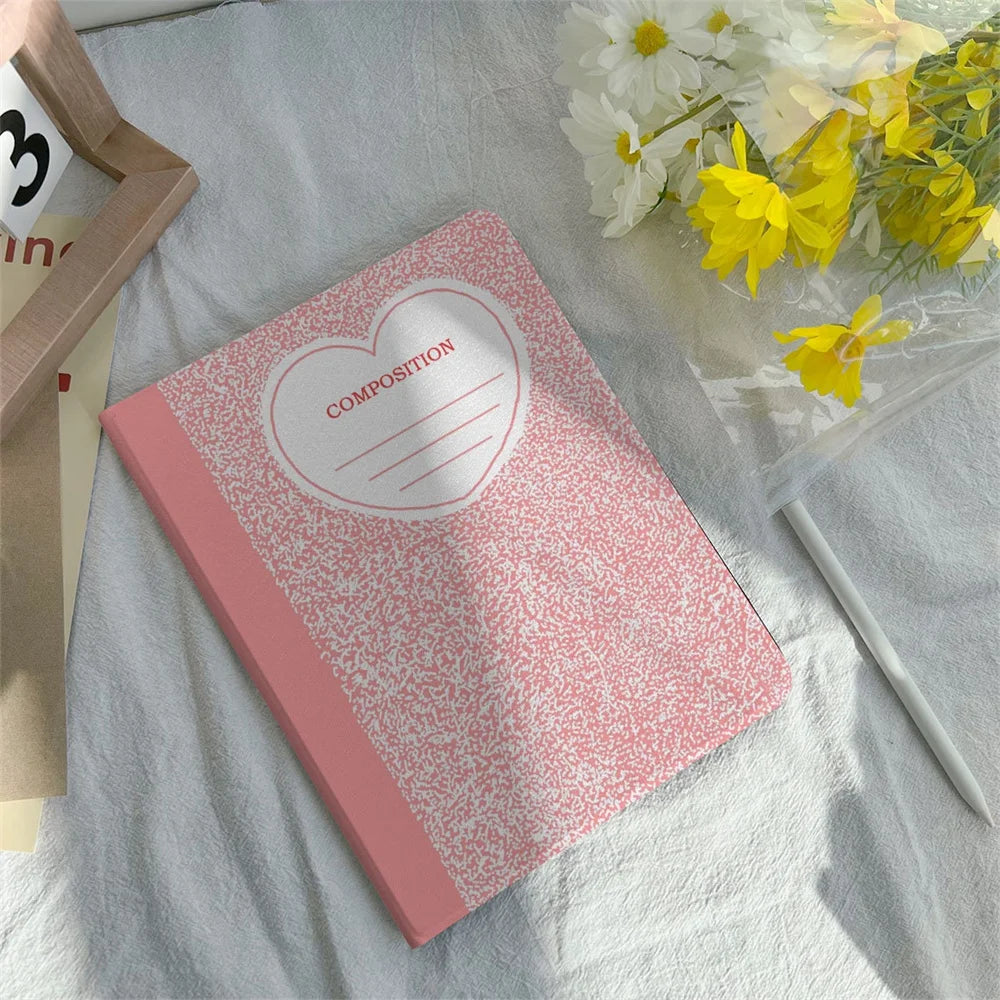 Pink composition notebook with a heart design and pencil holder lying on a white fabric.
