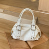 A white soft PU leather shoulder bag with silver rivet accents resting on a wooden surface.