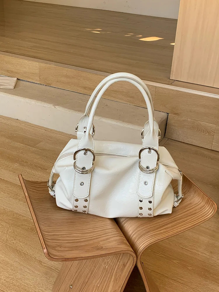 A white soft PU leather shoulder bag with silver rivet accents resting on a wooden surface.