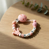 A pink silicone baby pacifier clip bracelet featuring a custom name and a small pink bead.